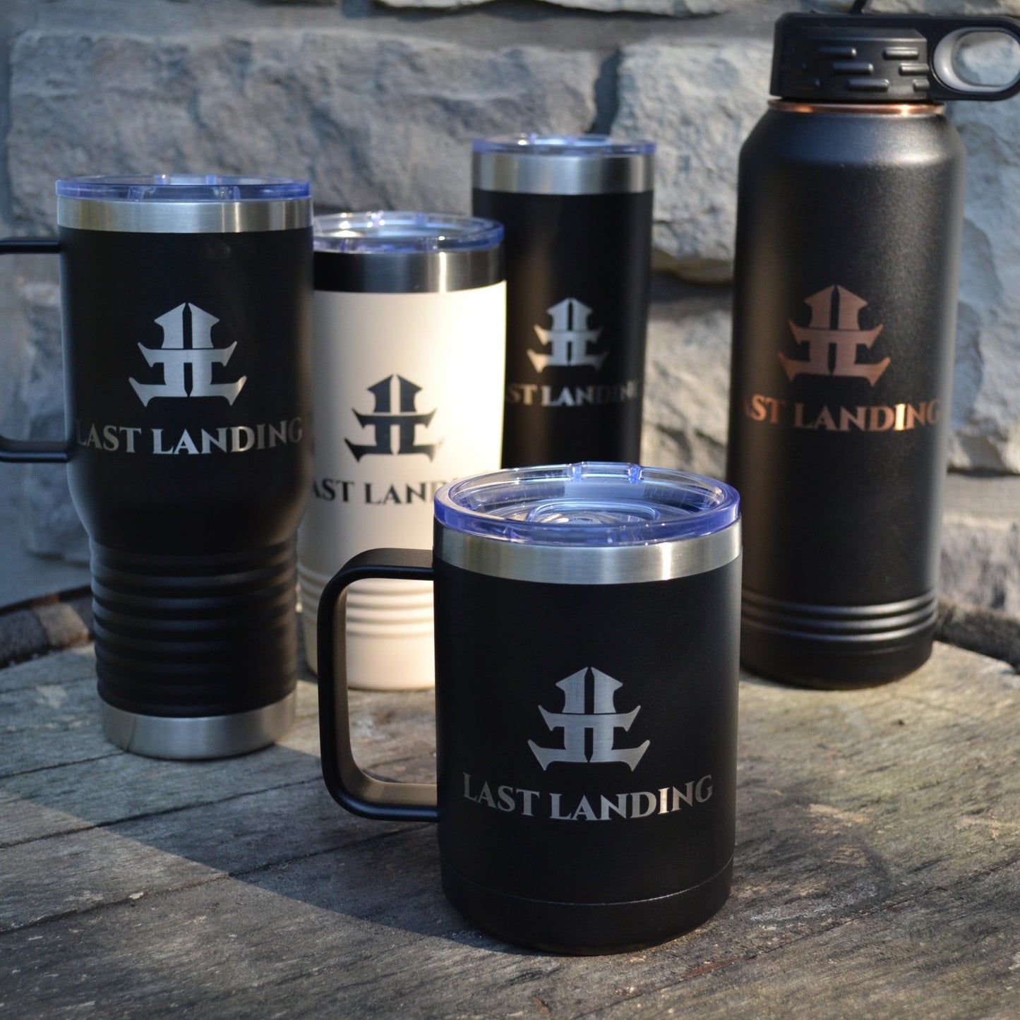 Set of black insulated mugs and bottles with 'Last Landing' branding on a stone surface.