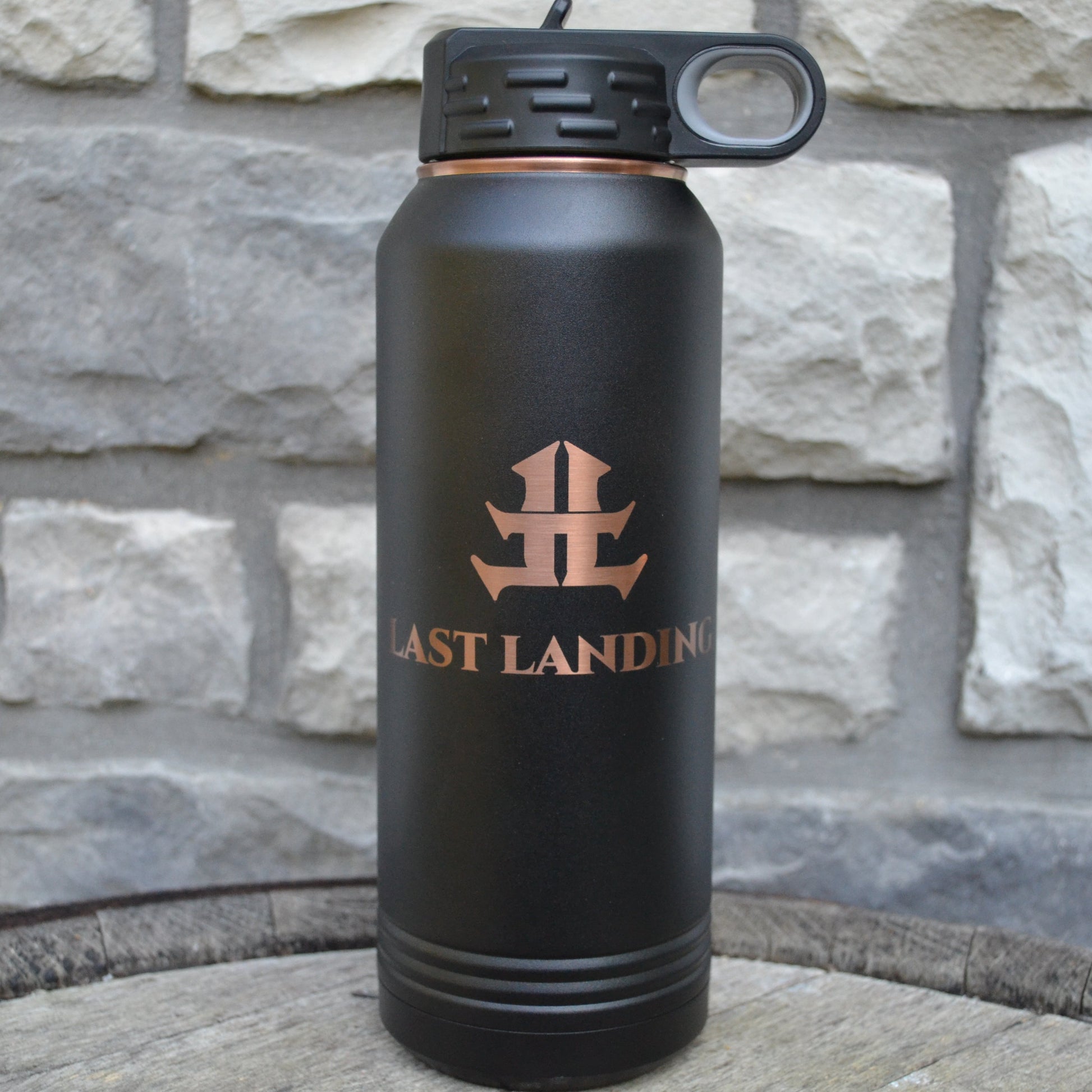 Black water bottle with 'Last Landing' logo against a stone wall background