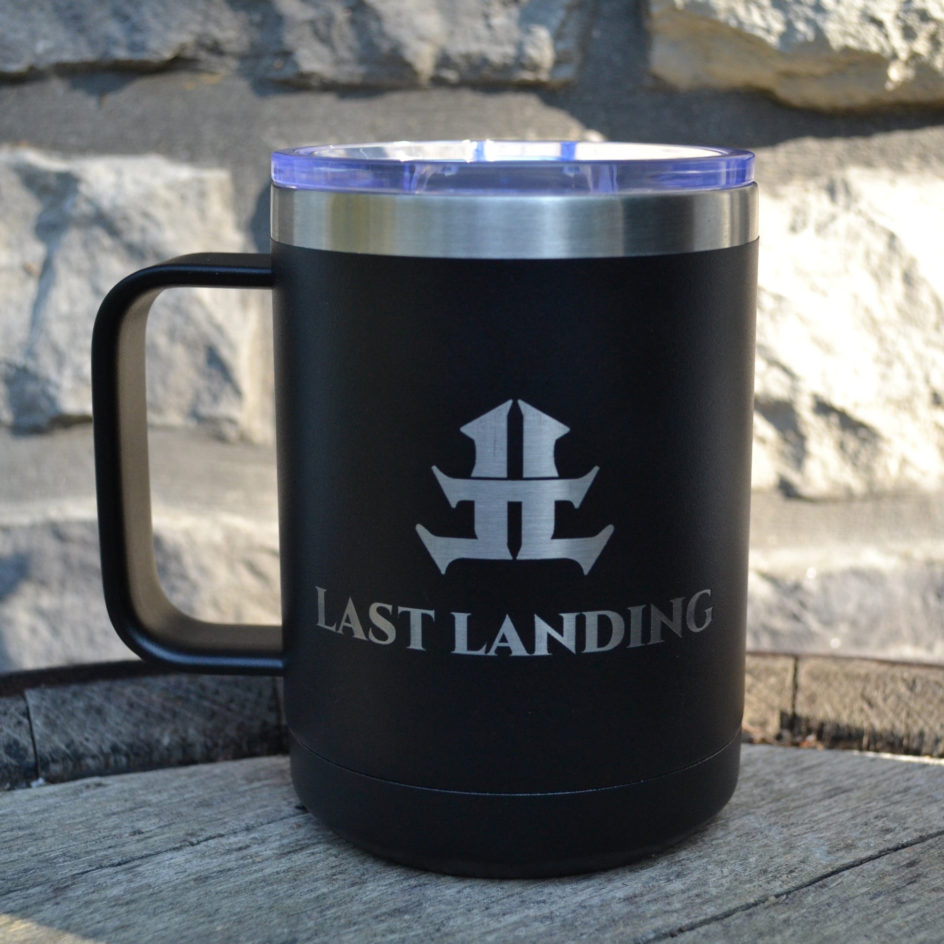 Black travel mug with 'Last Landing' logo on a stone surface and stone wall background