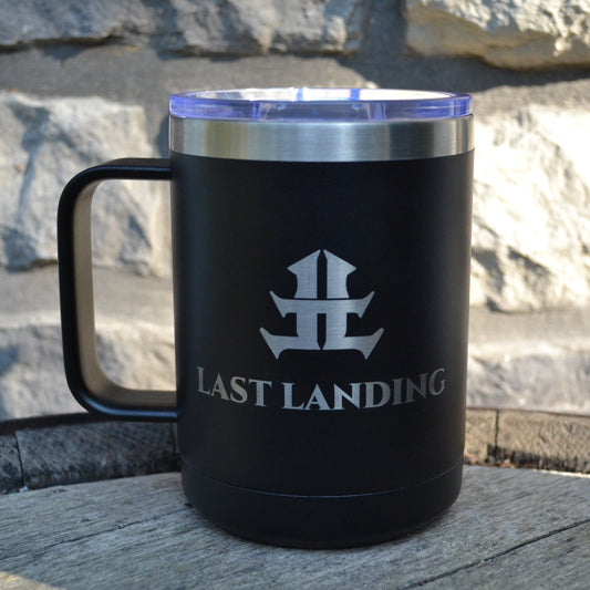 Black travel mug with 'Last Landing' logo on a stone surface and stone wall background