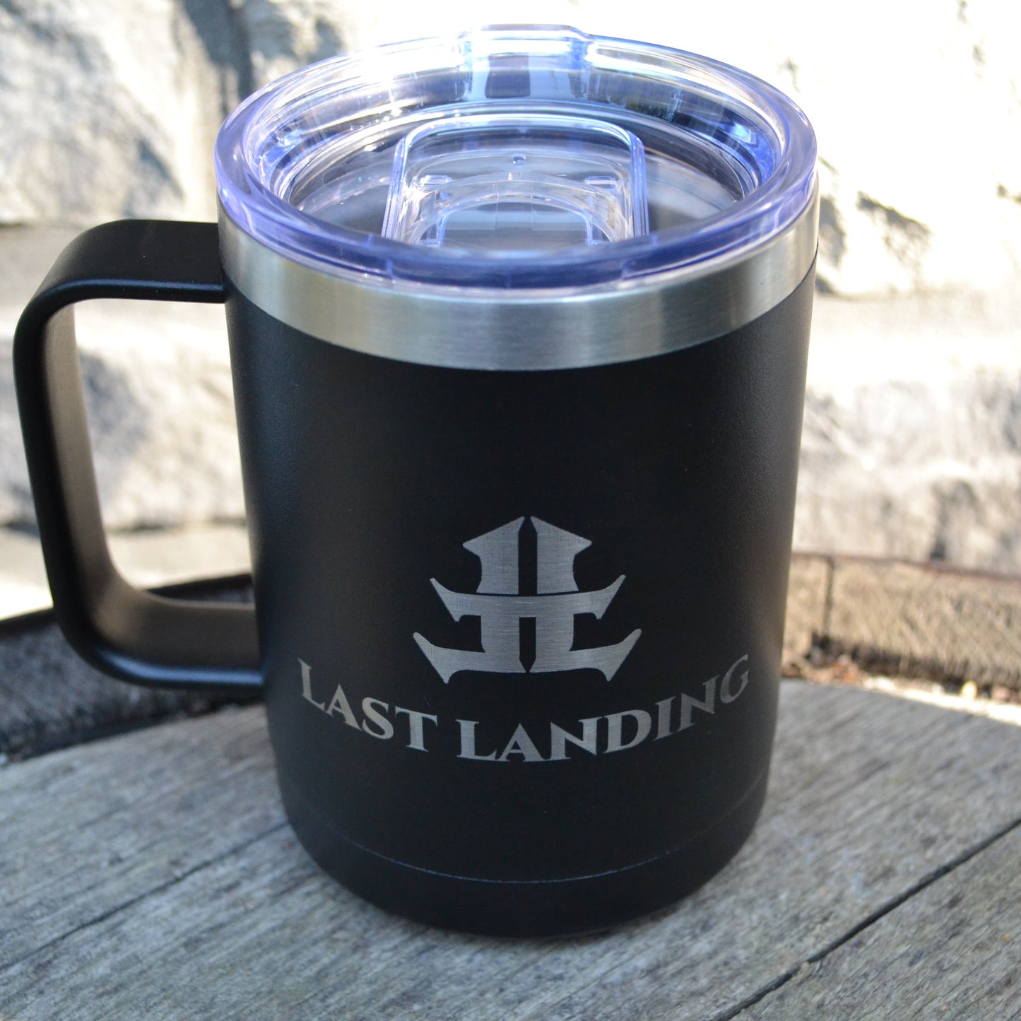 Black insulated mug with 'Last Landing' logo on a stone surface