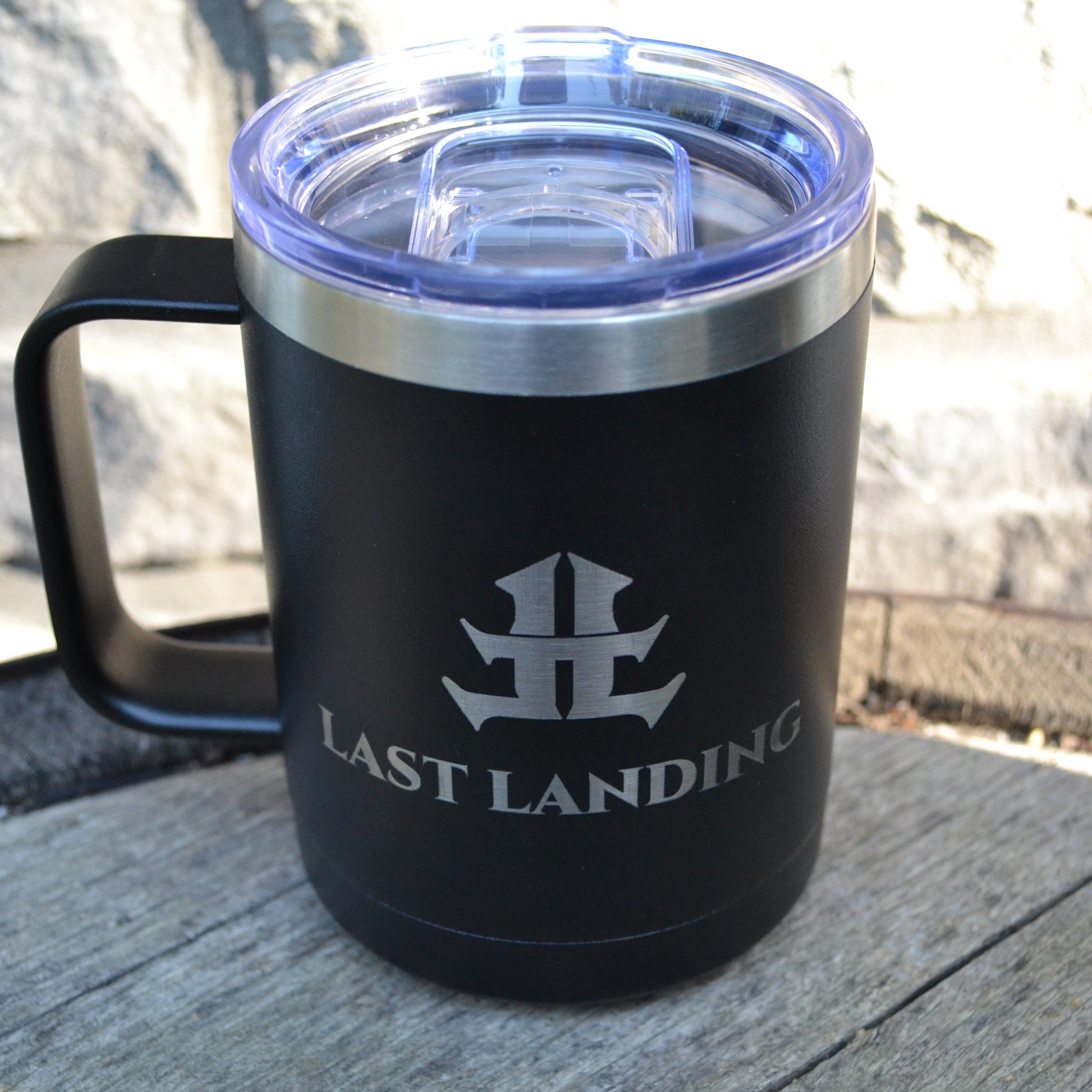 Black insulated mug with 'Last Landing' logo on a stone surface